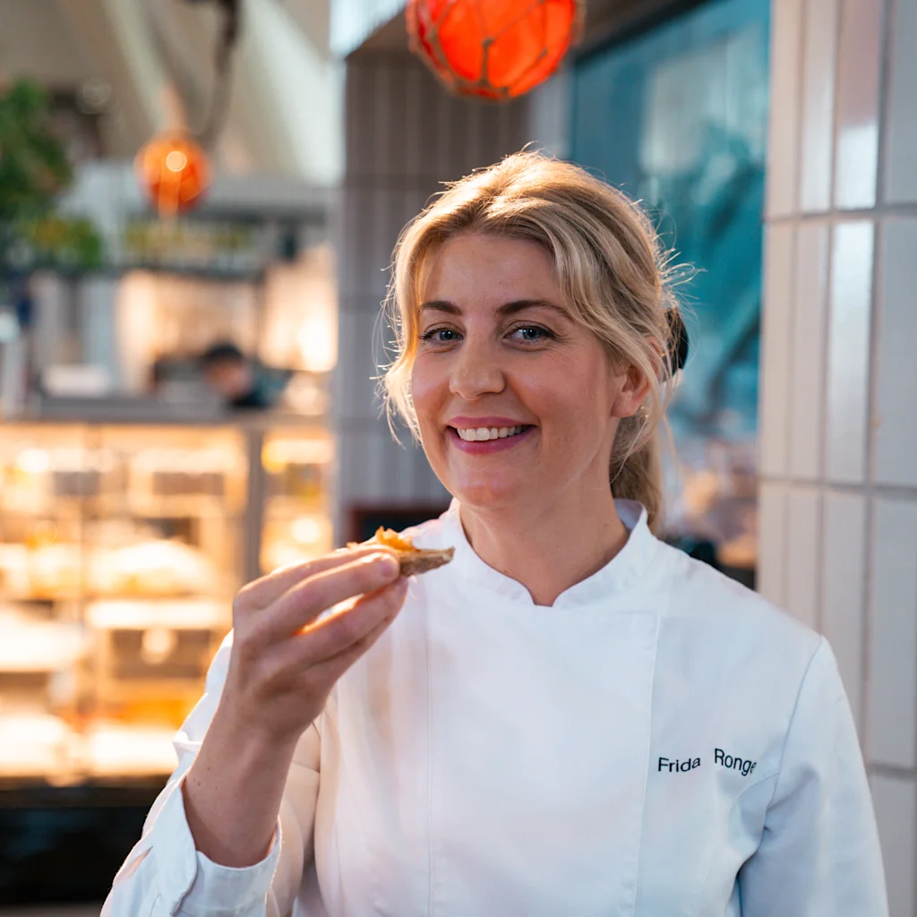A chef in a white coat named "Frida Ronge" enjoys a bite in a warmly lit restaurant, under hanging decorative orange spheres.
