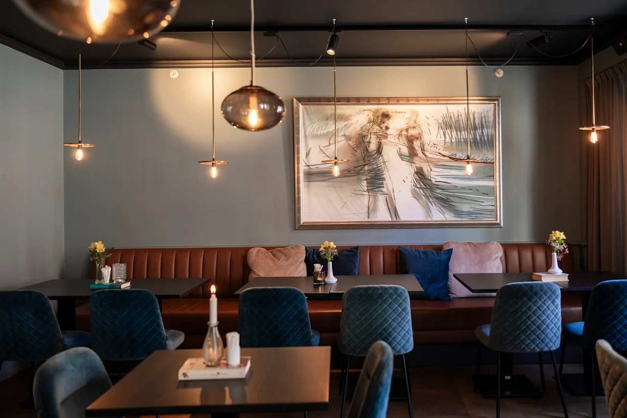 Moderna kaféets interiör med lång brun lädersoffa, teal-färgade stolar, träbord och hängande pendellampor. En stor abstrakt målning pryder väggen och skapar en mysig stämning.