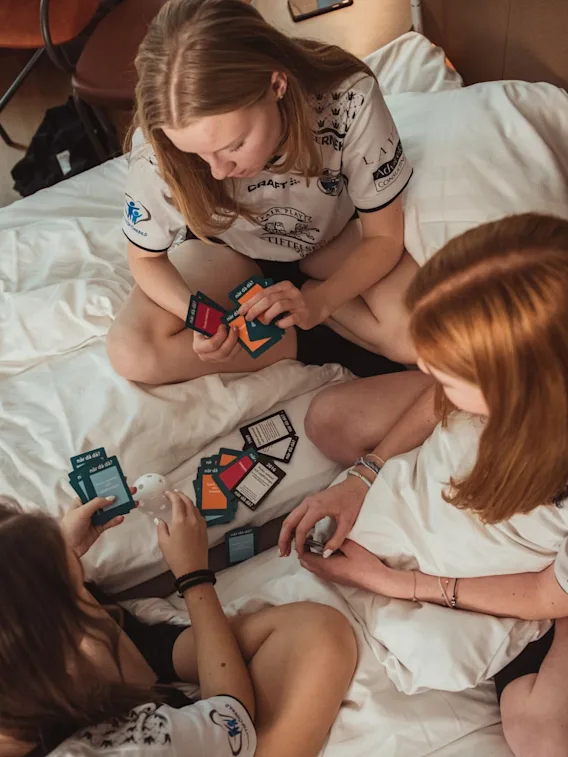 Tre unge idrettsutøvere sitter på en hotellseng og spiller et kortspill sammen.