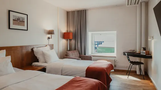 Viihtyisä hotellihuone, jossa kaksi erillistä sänkyä, työpöytä ja ikkuna, josta näkyy teollisuusalue.