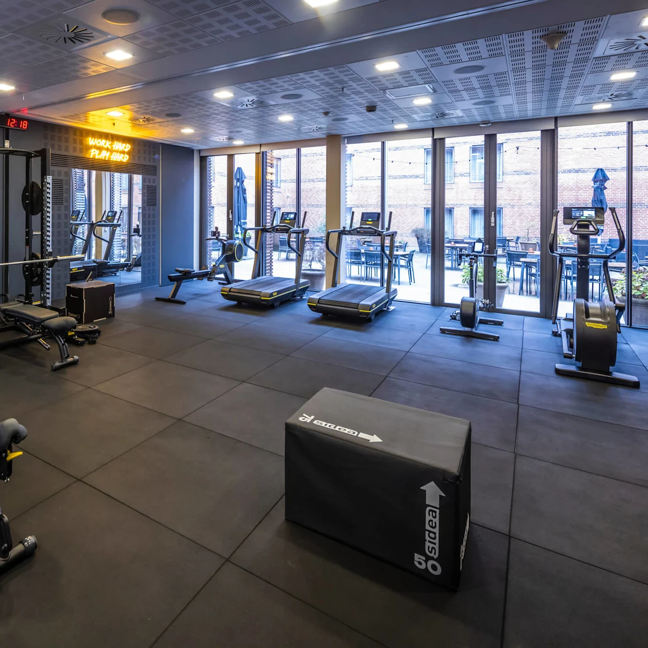 A modern gym with equipment such as dumbbells, a bench press, treadmills, and rowing machines. The room features rubber flooring and large windows allowing natural light