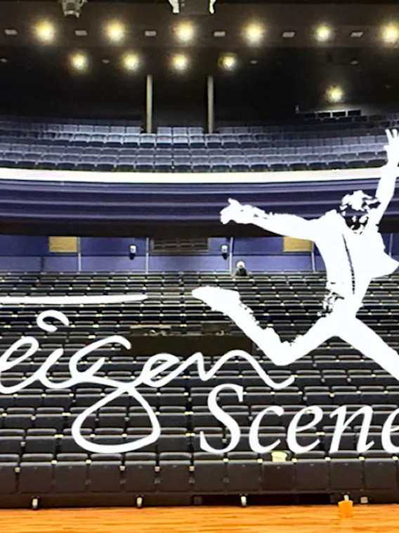 En person hopper på en scene i et stort auditorium med rader av seter. Ordene Teigen Scene vises.