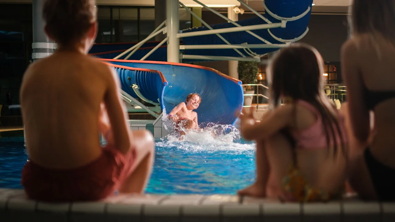 Children watching a boy splashing into an indoor pool from a water slide