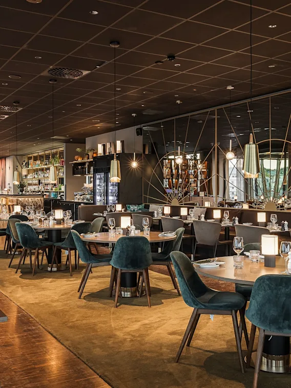 Elegant hotellrestaurang med mörkt tak, varm belysning och gröna sammetsstolar runt träbord.