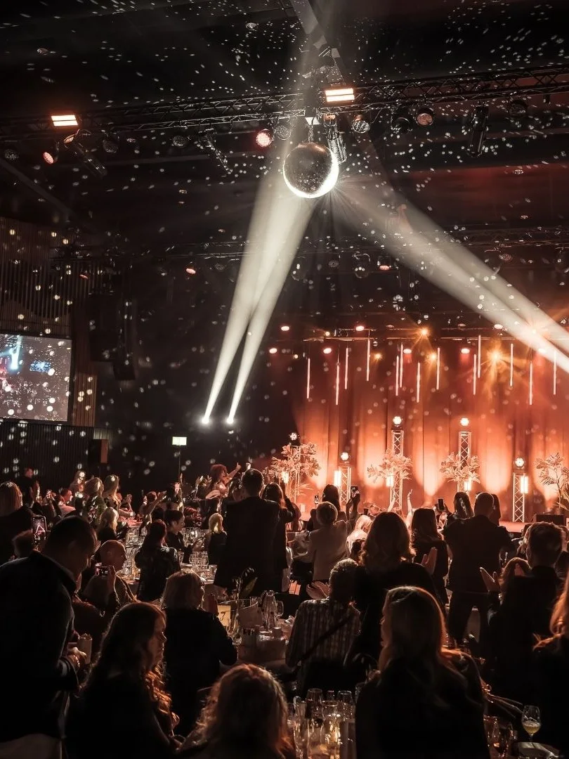 En publik tittar på en scenföreställning med starka ljus och konfetti. Runda bord fyller den svagt upplysta lokalen, medan stora skärmar visar evenemanget på sidorna.