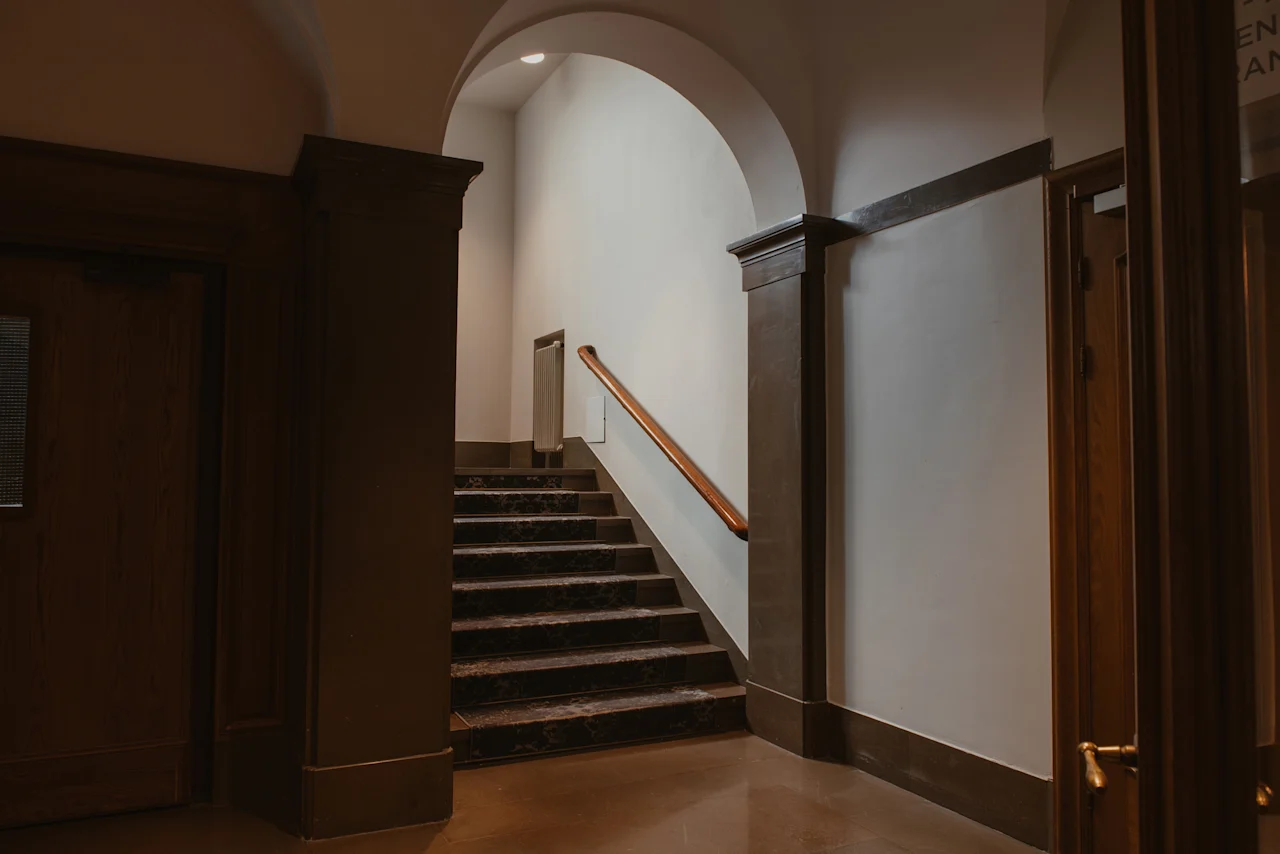 En storslagen trappa med mörka steg och ett träräcke som leder upp till en välvd hall på ett hotell.