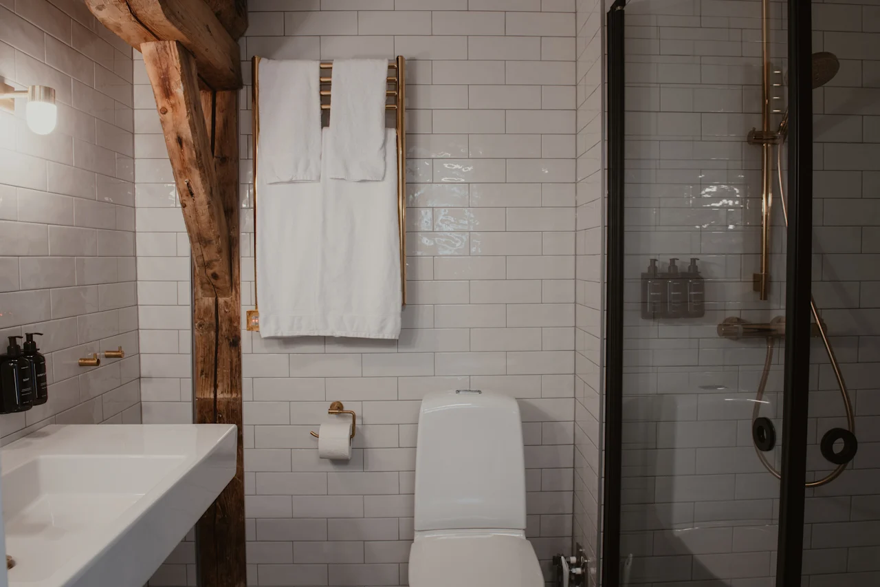 Et moderne badeværelse med hvide fliser, håndvask, toilet og bruser. Hvide håndklæder hænger på en gylden håndklædetørrer, og en rustik træbjælke tilføjer karakter.