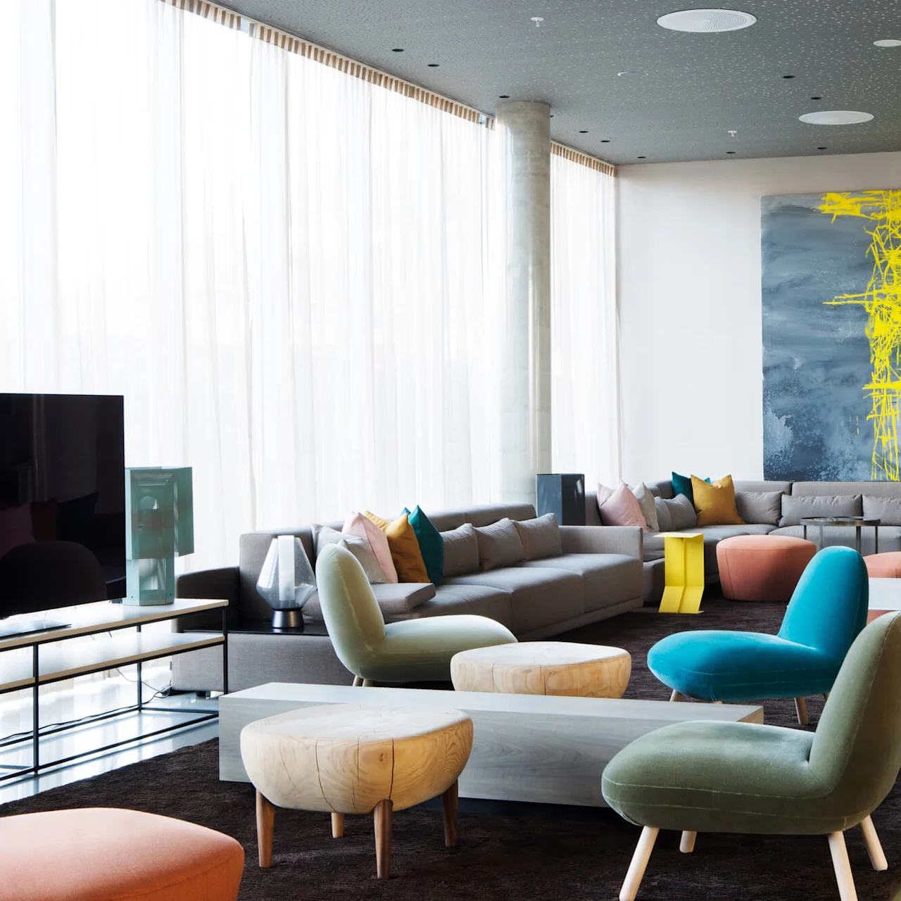 A spacious living room features modern furniture, including a sectional sofa with colorful pillows, armchairs, and a coffee table; set against floor-to-ceiling windows and abstract wall art.