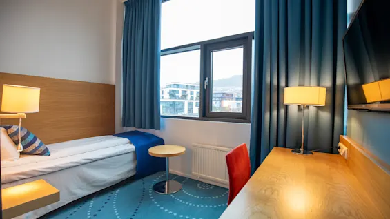 Et koselig hotellrom med enkeltseng, trepult og lampe. Stort vindu viser byutsikt, blå gardiner og teppe. Rød stol gir kontrast.