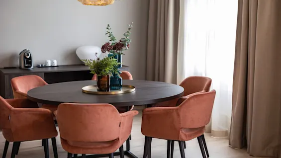 Et rundt træbord med blomster i midten er omgivet af lyserøde stole. En tekstureret pendellampe hænger over. Neutrale gardiner og et sidebord med en kaffemaskine fuldender rummet.