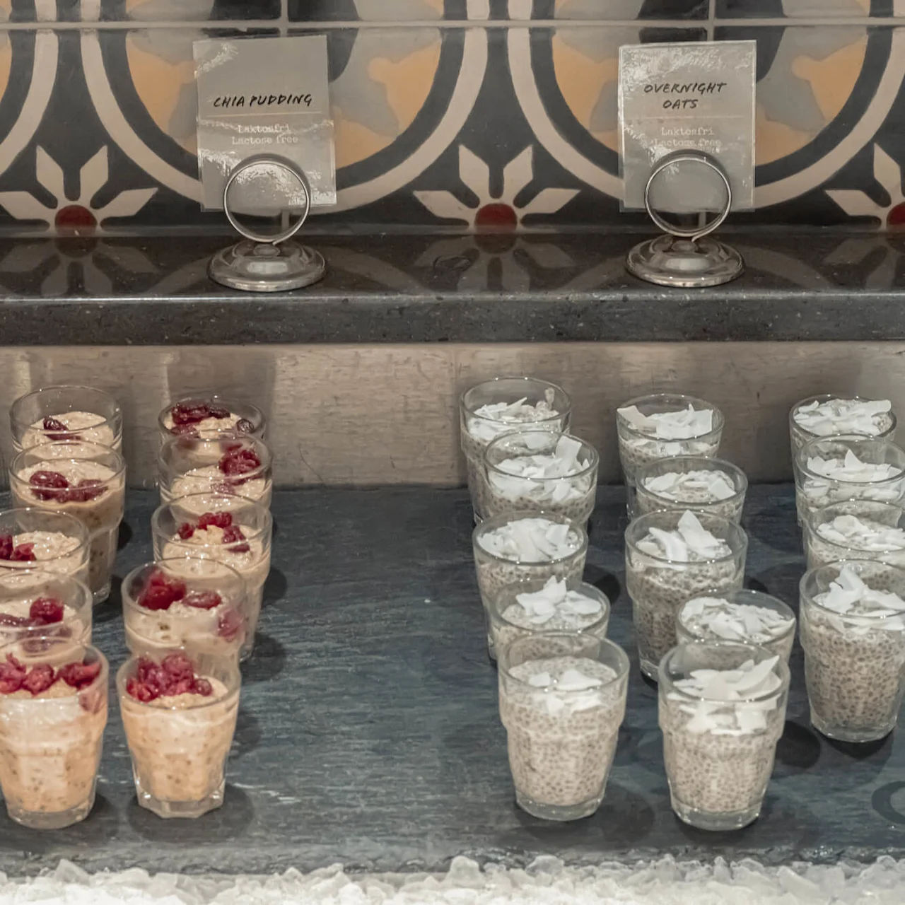 Rekker med chia-pudding og overnattet havre i glass, merket og servert på et brett med is, mot en dekorativ flisbakgrunn.