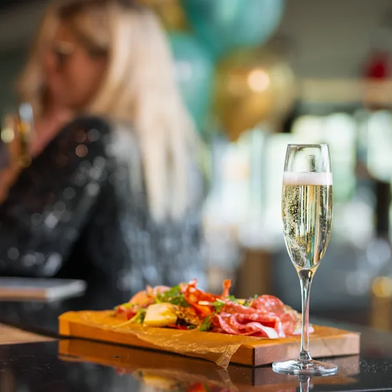 Champagneglass på disk, ledsaget av et charcuteriebrett toppet med kjøtt, grønnsaker og urter. Uklart bakgrunn avslører en person som holder et lignende glass i en festlig setting.