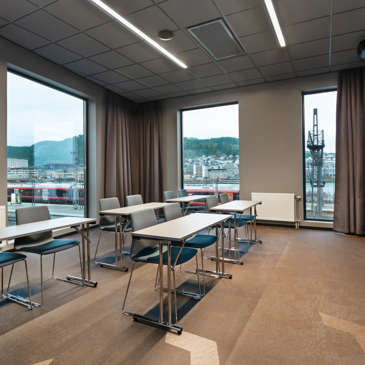 Bord og stoler er arrangert i rader mot en veggmontert skjerm i et moderne konferanserom med store vinduer som viser en byutsikt og åser utenfor.