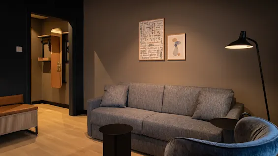 A modern hotel room with a grey sofa, armchair, and floor lamp. Wall art features a word cloud including STEINKJER, leikjaill, tullball.