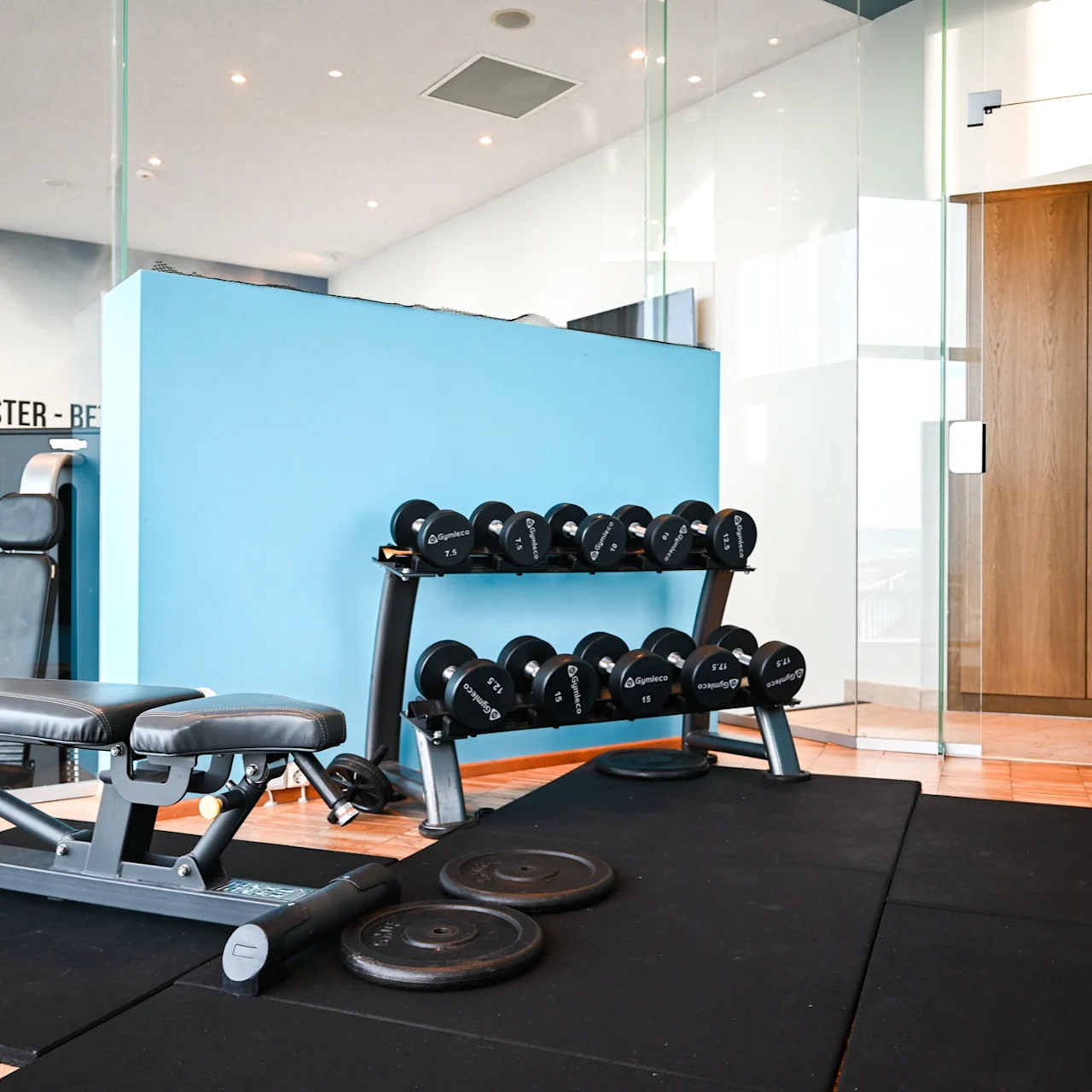 Weight bench and dumbbells are set up in a gym. Three weight plates lie on the floor. Text on the wall says "STRONGER - FASTER - BETTER."