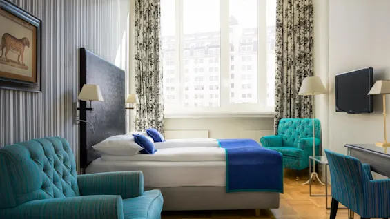 Et pent ordnet hotellrom med en seng, lenestoler, skrivebord og TV, belyst av naturlig lys fra store vinduer.