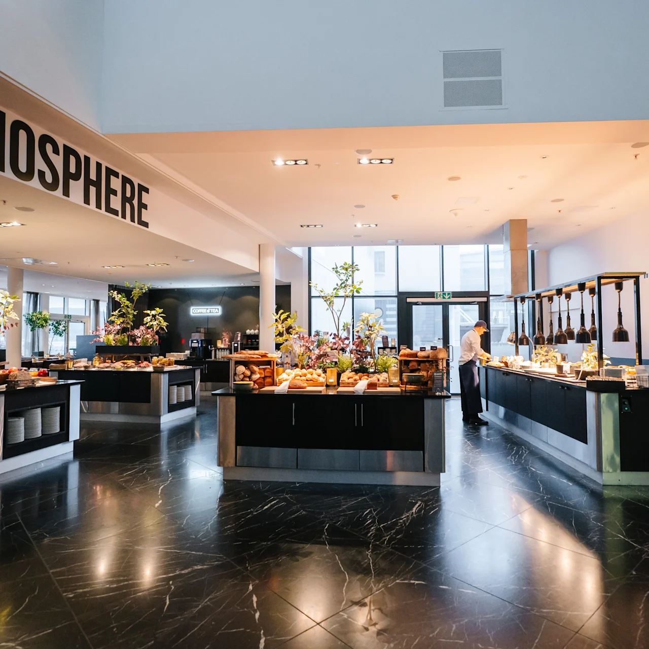 A spacious buffet area features tables filled with assorted food under soft lighting. Text on walls reads "ATMOSPHERE" and "LAUGHTER IS BRIGHTEST WHERE FOOD IS BEST." Staff are arranging items.