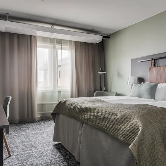 Ett modernt hotellrum med en bekväm säng, ett skrivbord och ett fönster med gardiner, som erbjuder en avkopplande vistelse.