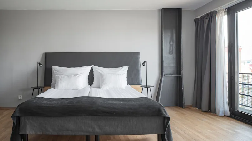 A neatly made double bed with white pillows and dark grey bedding sits in a modern bedroom with wooden floors and minimalist decor, adjacent to a window with grey curtains.