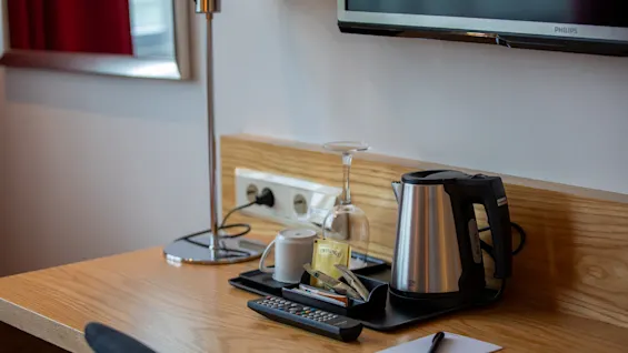 En vannkoker står på et trebord, omgitt av en kopp, et glass og teposer merket "Twinings". En fjernkontroll, lampe og en notisblokk med penn er i nærheten.