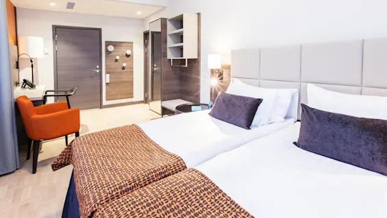 Ett hotellrum med två enkelsängar, täckta med vita sängkläder och mönstrade kastar, står i en modern miljö med stol, skrivbord och garderob mot ljusa väggar.