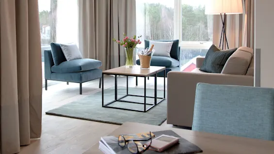 Stue med moderne sofa og stoler, plassert rundt et trebord med blomster. Gulv-til-tak-vinduer viser en skogsutsikt, som skaper en rolig atmosfære.