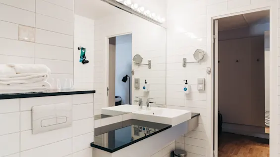 Bright hotel bathroom with white tiles, a large mirror, sink, and towels.