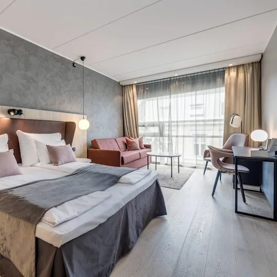 En pent dobbeltseng med myke puter og mørkt sengeteppe står i et moderne hotellrom. I nærheten er det en sofa, et skrivebord og et stort vindu som skaper en romslig, lys atmosfære.