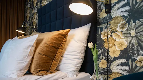 A bed with white and gold pillows rests beneath a dim wall-mounted lamp in a room featuring tropical floral wallpaper and a small table with a vase holding a single tulip.