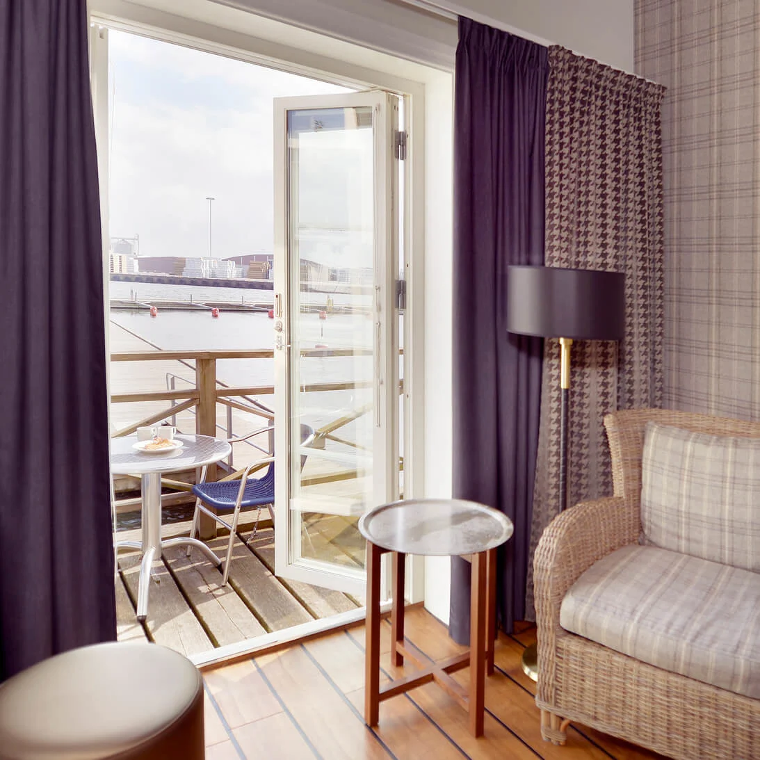 A cozy hotel room with a wicker chair and bedside lamp, opening to a balcony overlooking water.