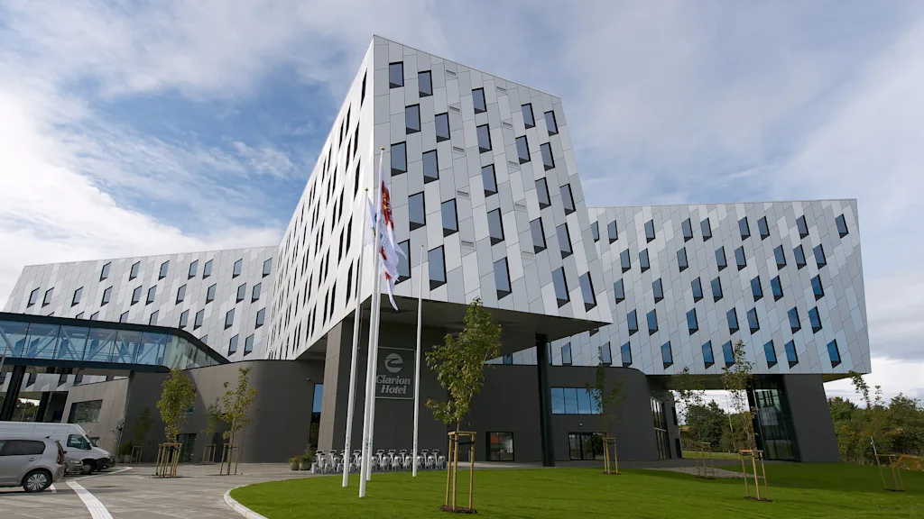 En moderne, geometrisk udformet bygning med skrånende vægge og mange vinduer står under en blå himmel. Ved indgangen står "Clarion Hotel" og flere flag vifter i et haveområde.