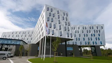 En moderne, geometrisk utformet bygning med skrå vegger og mange vinduer står under en blå himmel. Ved inngangen står det "Clarion Hotel" og flere flagg vaier i et hageområde.