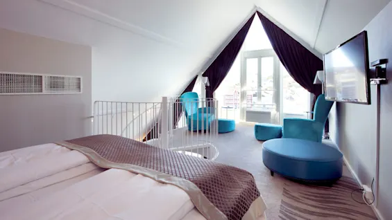 A neatly arranged hotel room with a large bed, blue accents, and an open loft area under a sloping ceiling with a window providing natural light.