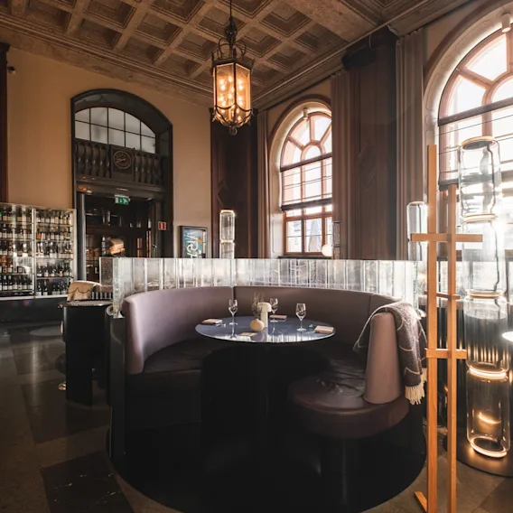 An elegant hotel lounge or restaurant area with a cozy booth seating, a set table, and large arched windows letting in bright sunlight.
