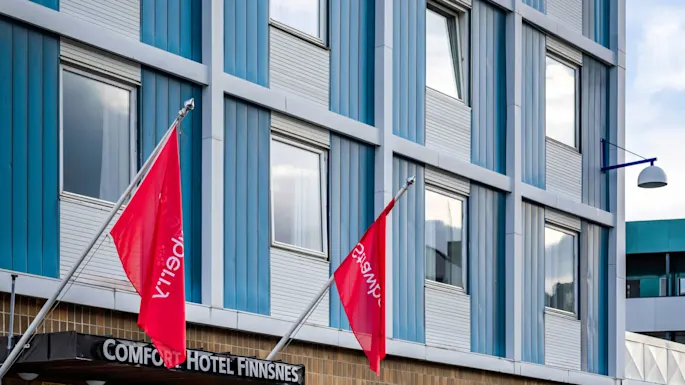 A hotel entrance with "COMFORT HOTEL FINNSNES" sign, flanked by two red flags and white chairs. Surrounding glass windows reflect the cloudy sky.