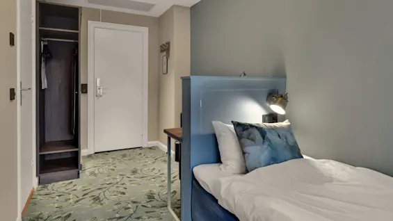En säng med en blå sänggavel, vitt lakan. Öppet skåp med kläder, mattat golv. I ett hotellrum.