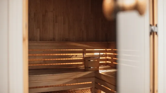 Trebenk står tom, bøtte plassert på den, inne i en varmt belyst badstue med panelvegger og mønstret gulv, delvis synlig gjennom en åpen dør.