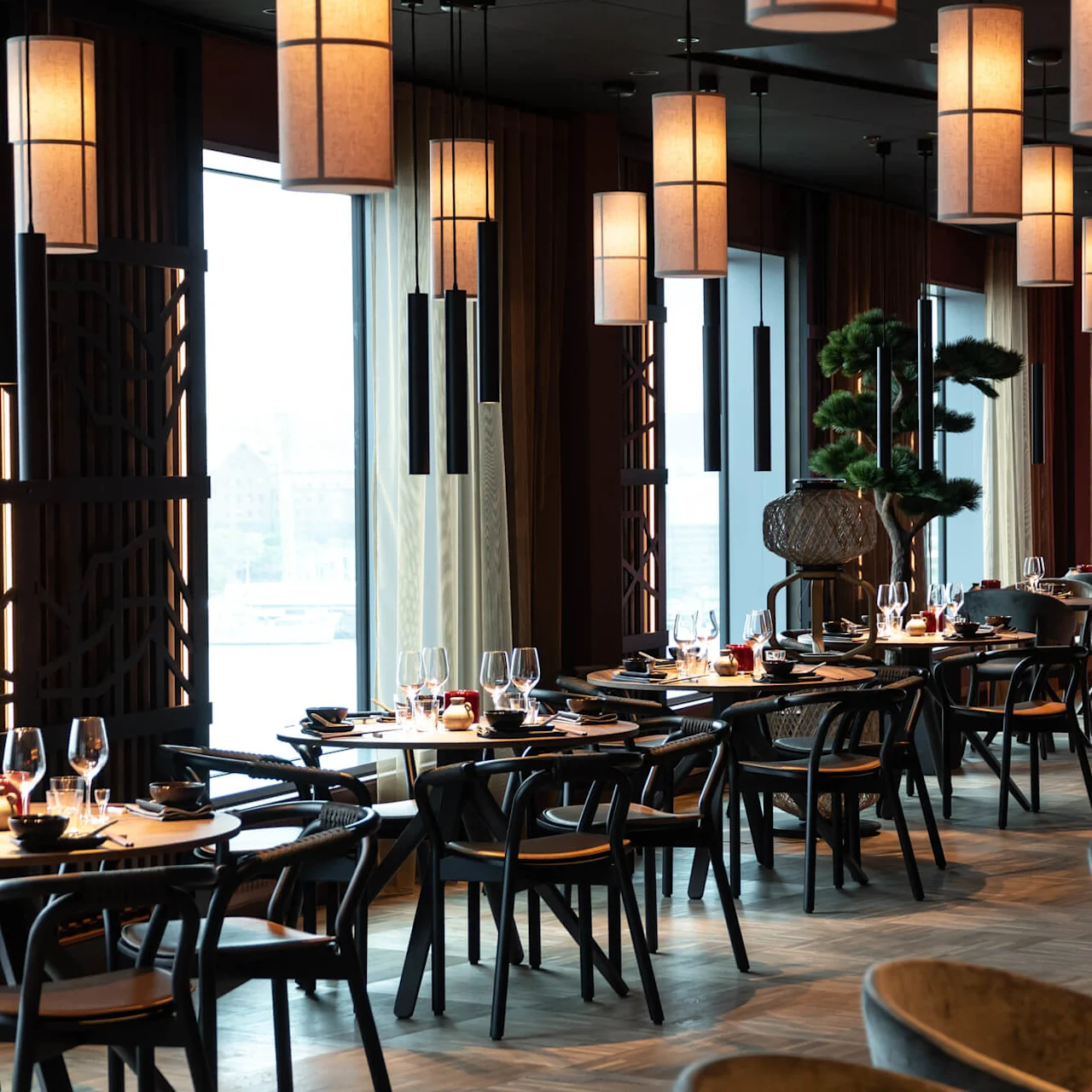 Restaurangbord är elegant dukade med vinglas och bestick, omgivna av modern inredning och varma hängande lampor. Stora fönster ger naturligt ljus medan dekorativa träd förhöjer miljön.