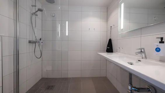 Showerhead affixed to tiled wall with glass partition in a modern bathroom, adjacent to a long sink with a mirror and soap dispenser. Gray floor tiles and overhead lighting.