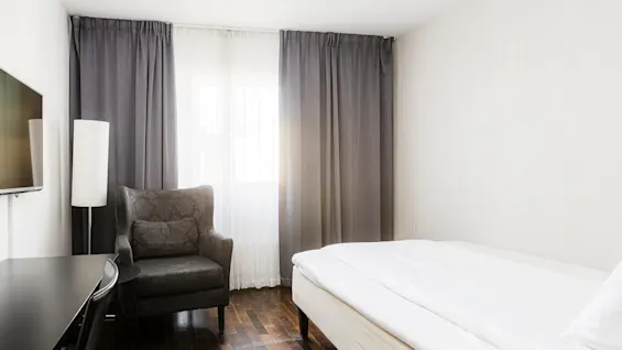 En mörkgrå fåtölj vätter mot ett fönster täckt av tunna och ogenomskinliga gardiner i ett minimalistiskt sovrum, kompletterat med en säng och en väggmonterad TV.