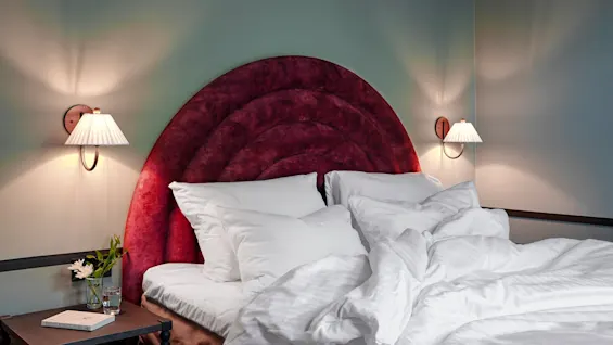 A bed with white rumpled sheets and pillows sits beneath a red, plush headboard. Wall-mounted lamps illuminate the room, with a nightstand holding a book and a vase of flowers.