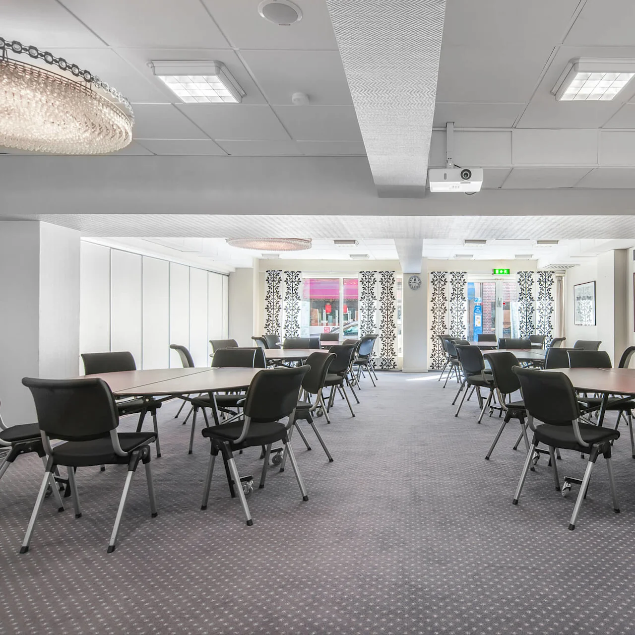 Bord och stolar är arrangerade i ett rymligt, väl upplyst konferensrum med mönstrade gardiner och konstverk på väggarna, vilket skapar en professionell mötesmiljö under modern belysning.
