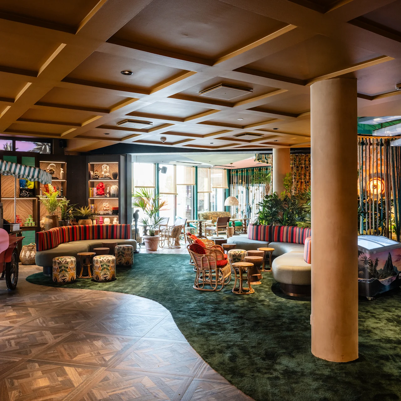 Colorful interior features eclectic furniture arranged in a cozy seating area, with potted plants and warm lighting, creating a vibrant and inviting atmosphere. Wooden flooring contrasts with green carpet sections.