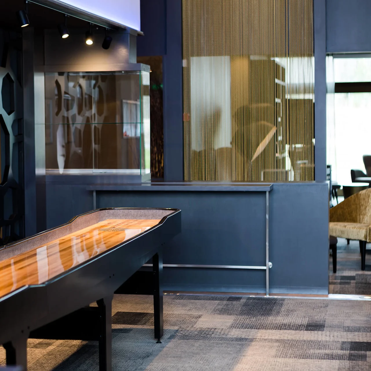 A wooden shuffleboard table stands in a modern room with a geometric-patterned wall. A red mannequin torso is blurred in the foreground.