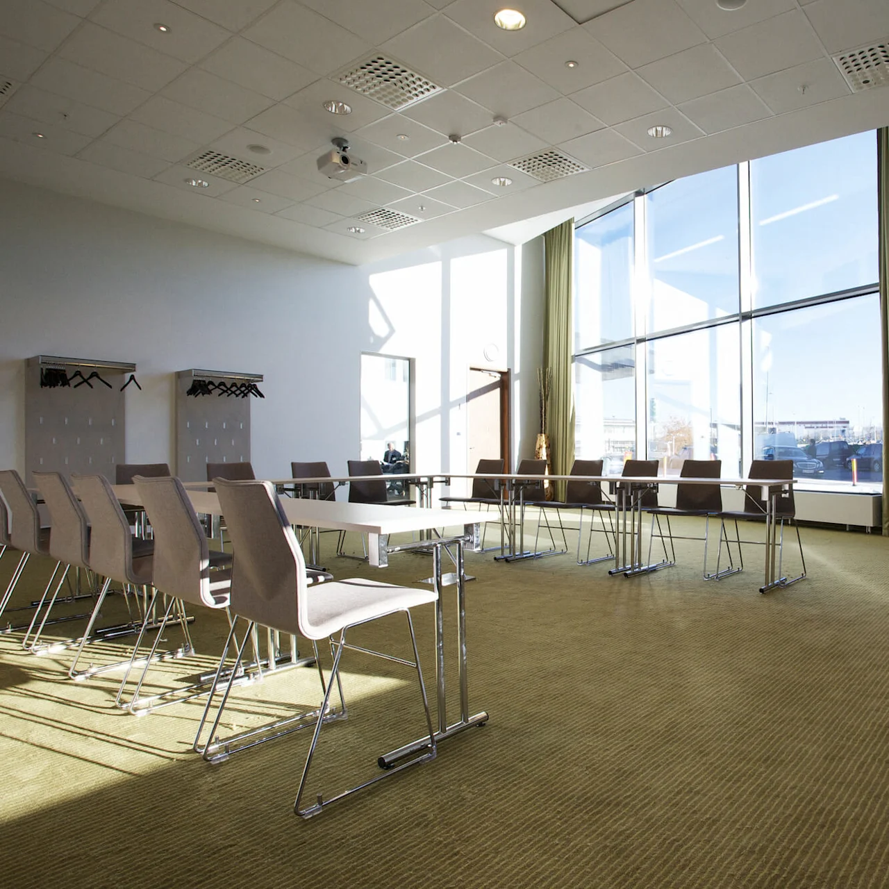 Wide conference room contains arranged chairs and tables, illuminated by sunlight streaming through large windows with green curtains; coat racks hang on wall above textured carpet.