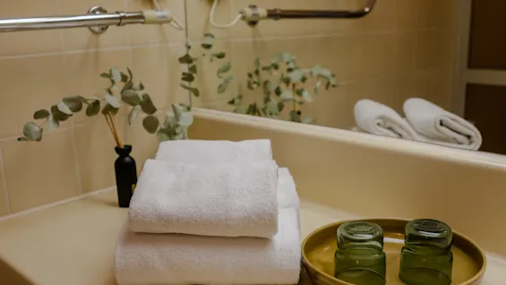 White towels stacked beside green glasses on a tray, surrounded by eucalyptus branches, placed in a bathroom with beige tiles and a mirror.