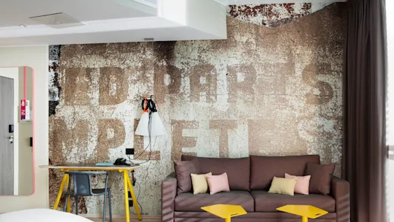 A cozy living area features a brown sofa with pastel pillows, yellow tables, and a grunge-textured wall with faded text: "TID • • • P R A • • • S COMPLETE”. Bright daylight filters through curtains.