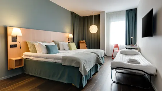 Et moderne hotellrom inneholder en stor seng og en mindre sammenleggbar seng, med stemningsbelysning og mørke tregulv. Et vindu med gardiner lyser opp rommet.