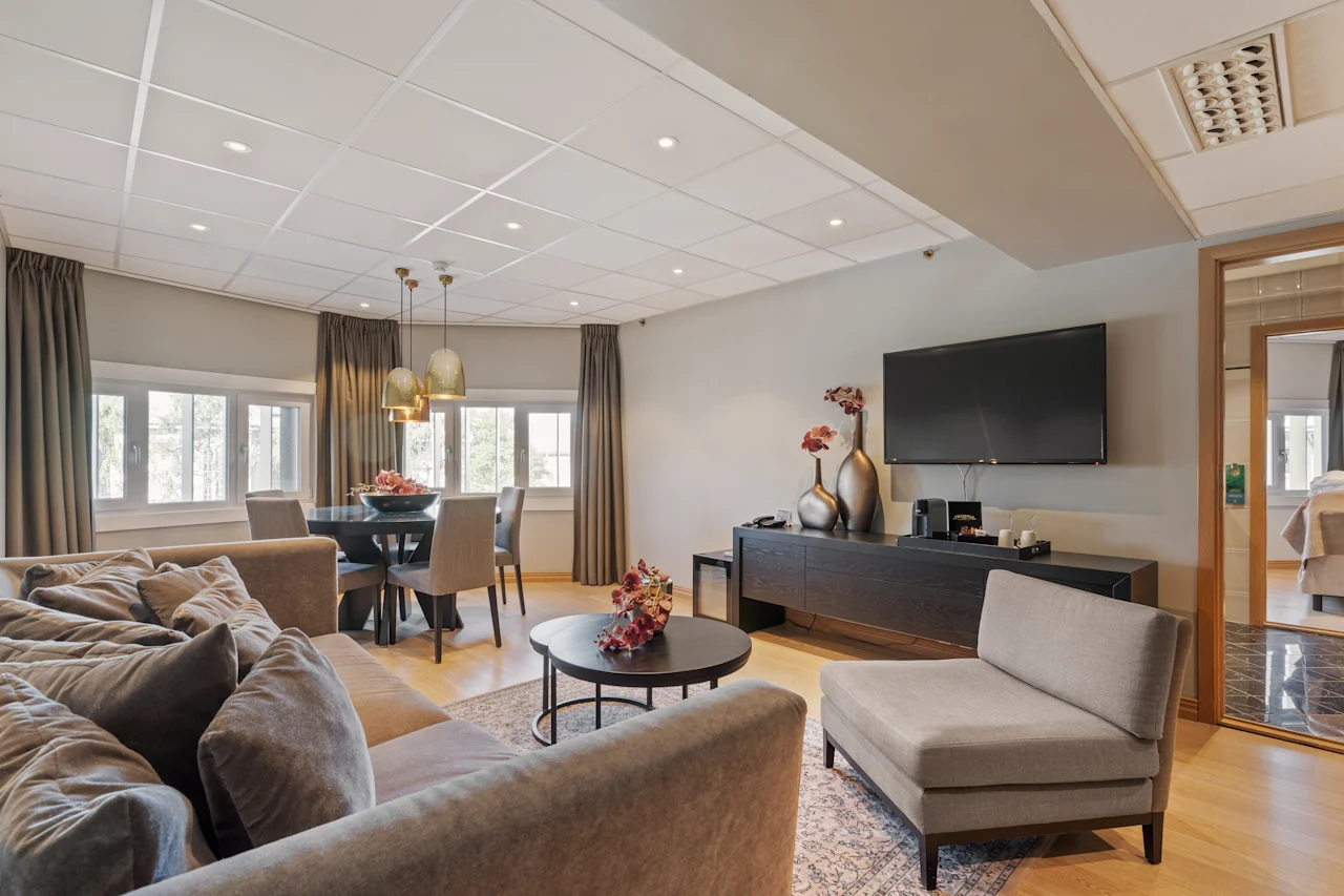 En stue med en grå sofa og stol står ved siden af et kaffebord med blomster; et spiseområde med vinduer og pendellamper er i nærheden, mens et TV hænger på væggen.