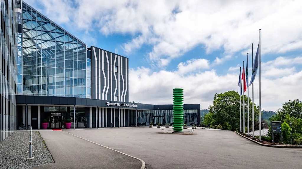 A modern hotel building displaying "QUALITY HOTEL EDVARD GRIEG" has glass facades with abstract patterns. Flagpoles and a green sculpture stand outside against a partly cloudy sky.